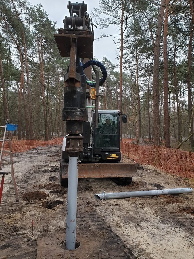 Excavadora instal·lant un pilot helicoïdal galvanitzat entre els pins del bosc de Bosland, Bèlgica